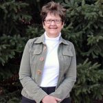 Diane Larsen, a women with short brown hair and glasses smiles, standing in front of pine tree