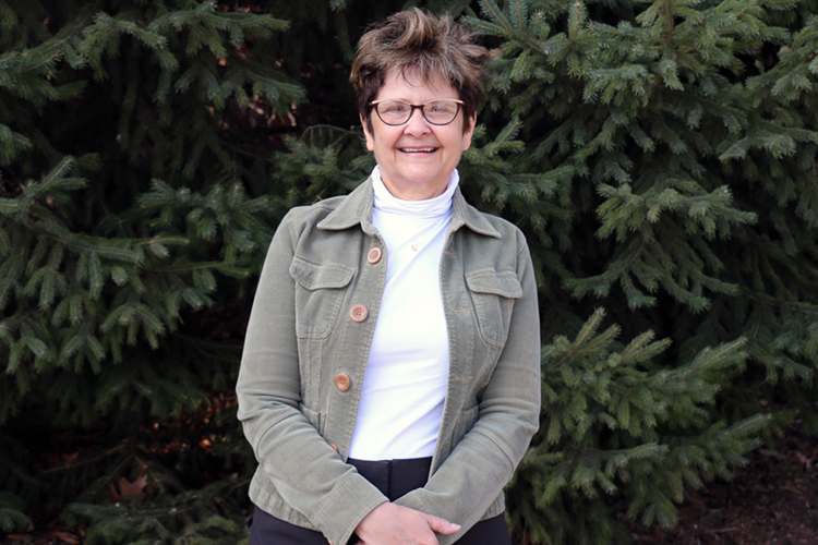 Diane Larsen, a women with short brown hair and glasses smiles, standing in front of pine tree