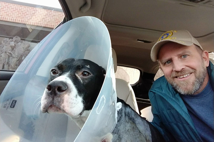 Man sits in car with his dog, Sarge, wearing a plastic head cone.