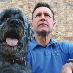 Man in blue shirt sits with his big black dog.