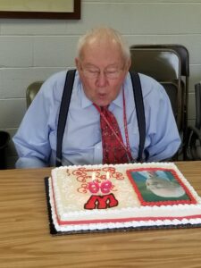 Bernard Easterday, wearing a blue shirt and red tie, blows out candles on a decorated cake with his photo. He looks joyful, celebrating a special occasion.