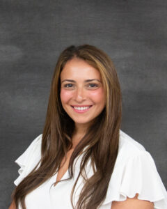 A long brown hair with highlights woman wearing a white blouse. Beatrice DeMarco