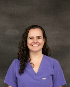 A log dark brown curly hair woman wearing a medium blue scrub top. Dani Labonte