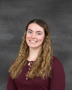 A long curly sandy brown hair wearing a long sleeve burgundy henley shirt. Elisse Friedman