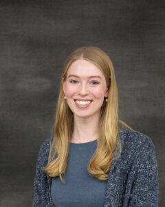 A long sandy blonde hair with grey blue sweater and top. Erika Mueller