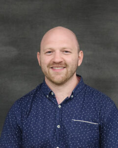 A bald man with low beard wearing a print navy button up shirt. Petter Burrows