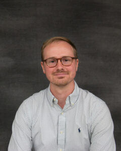A short sandy blonde hair main with glasses, wearing plaid gray button up long sleeve shirt. Samuel Hutchens
