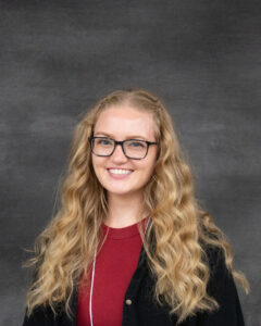 A long curly blonde hair woman with glasses, wearing a black sweater and red shirt. Sydney Worley