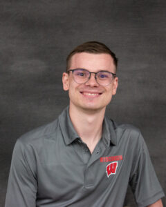 A man with short hair and glasses, wearing gray polo shirt with UW Wisconsin logo on left chest. Tanner Komlodi